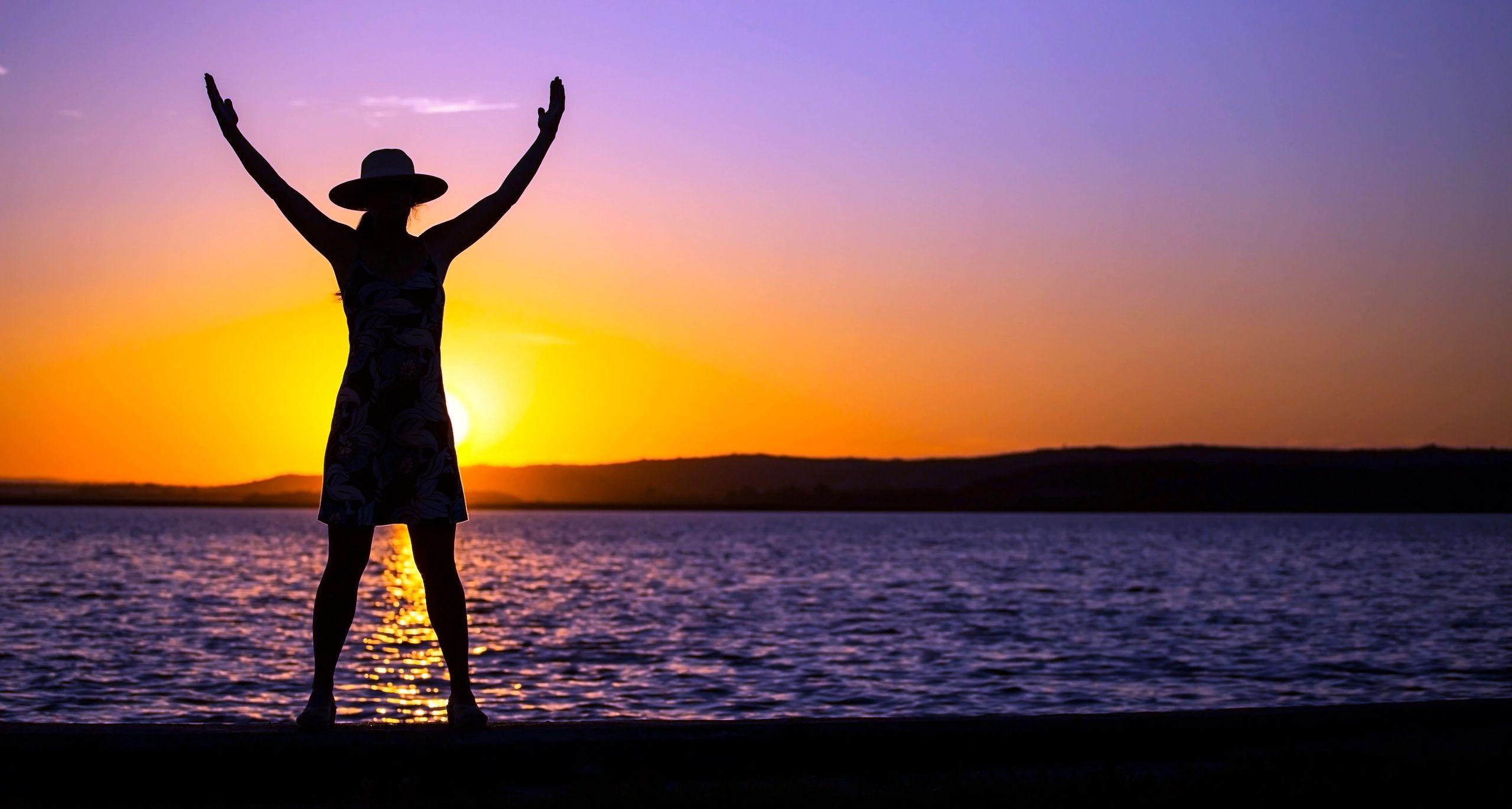 A woman raising both arms toward a purple and gold sunset over calm water